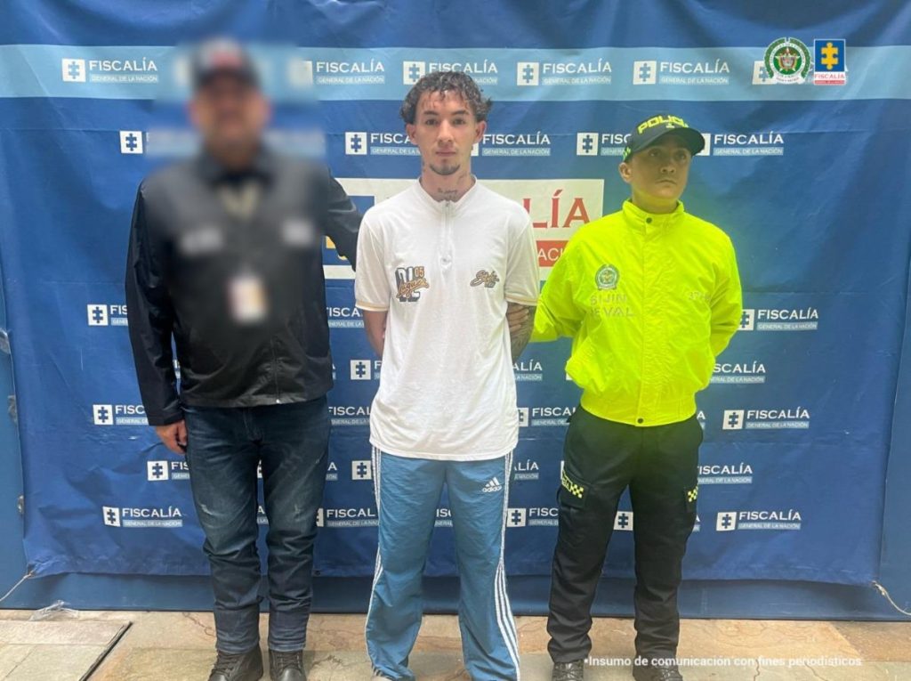 en la imagen se puede ver al capturado en medio de un funcionarios de la policía y de la fiscalia, detras de ellos se encuentra un baking de fiscalia