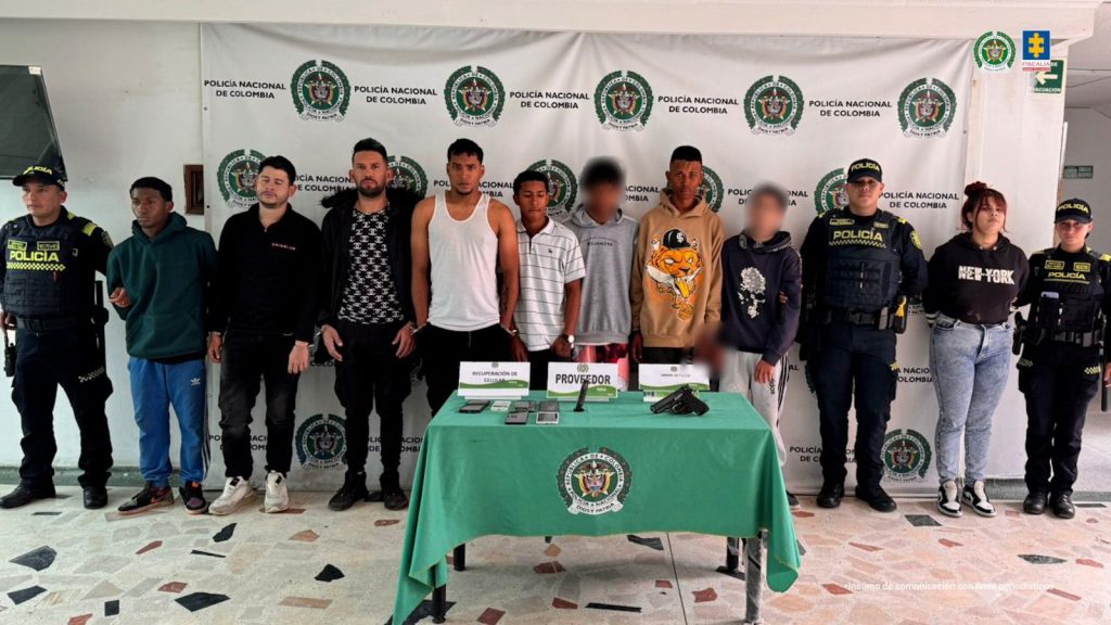 En la fotografía se ven los 9 capturados entre ellos una mujer, estas personas estan en medio de dos agentes de policia. Atra de ellos se encuentra un baking y delante hay una mesa con los objetos incautados, como celulares y armas de fuego y municiones
