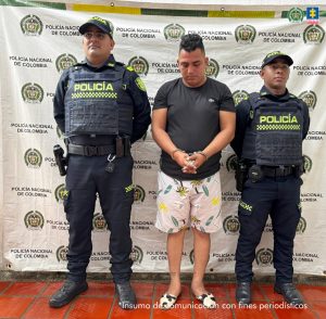 En la foto se ve un backing con logos de la policía. Allí aparece un hombre esposado en camiseta y pantaloneta y en cada lado del sujeto hay un Policía con su respectivo uniforme de color Azul.