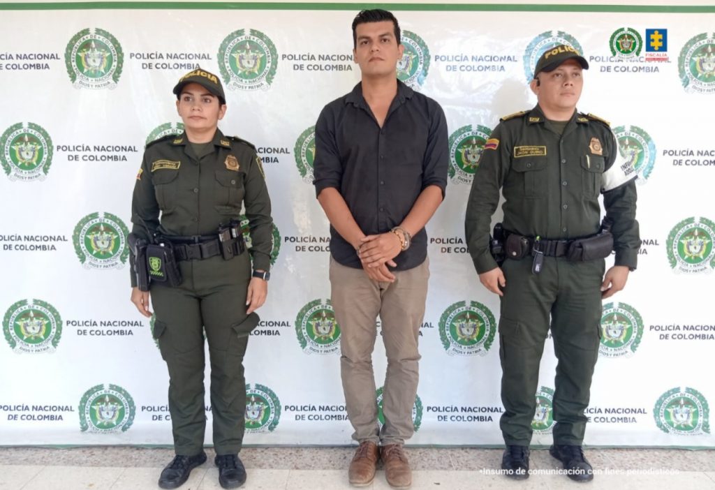 En la foto aparecen dos agentes de Policía a cada extremo del capturado. La persona viste una camisnegra y un pantalón caqui. Está esposado. De fondo hay un backing con logos de la Policía Nacional. Y en la parte derecha superior los logos de la Policía y de la Fiscalía General de la Nación.