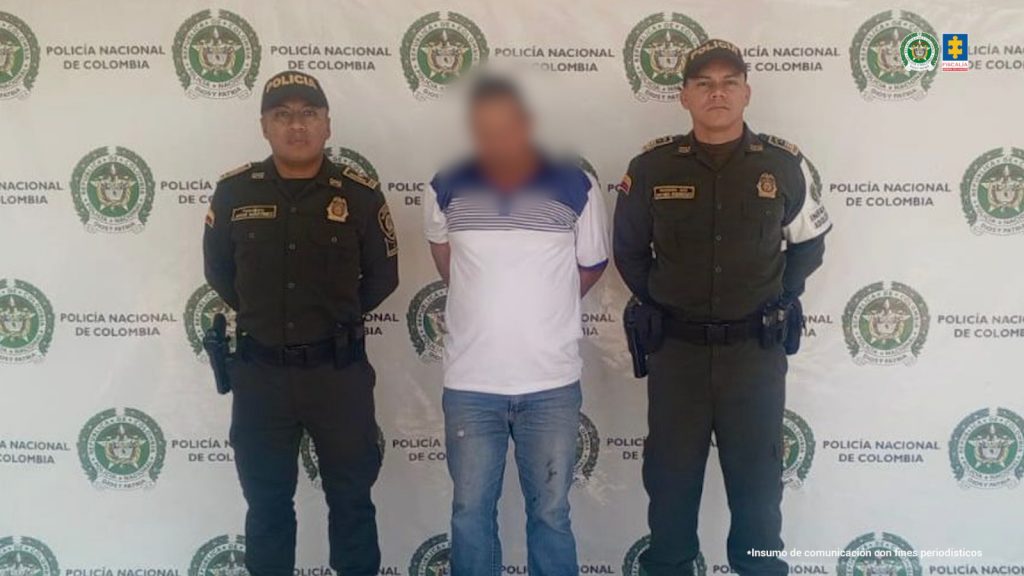 En la foto aparecen dos agentes de Policía a cada extremo del capturado. El señalado usa una camiseta blanca con rayas azules en la parte superior, está esposado y su rostro está tapado debido a que tiene parentesco con la que sería su víctima. De fondo hay un backing con logos de la Policía Nacional. Y en la parte derecha superior los logos de la Policía y de la Fiscalía General de la Nación.