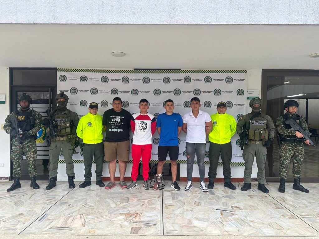 fotografia en donde se ven a los4 capturados en medios de dos agentes de la policia y dos del ejercito nacional, atras de ellos hay un baking de la policia