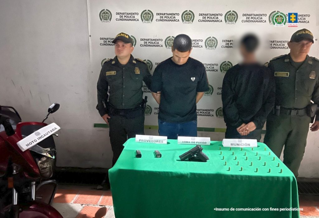 en la imagen se puede ver dos capturados, uno de ellos de espalda y difuminado por ser menor de edad, estas dos personas estan entre do agentes de policia, delante de ellos se encuentra una mesa con el material incautado y al lado de la mesa una moto. detras de todo se ve un bakin de la policia