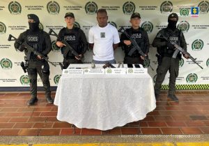 En la fotografía aparece de pie y con los brazos atrás Jhon Freddy Rivera Montaño, alias JF. Viste con camiseta de color blanco, manga corta y con estampado rojo en el pecho; asimismo una pantaloneta azul. A sus extremos hay uniformados de la Policía Nacional. Delante de ellos hay una mesa con mantel blanco y sobre ella hay unos documentos, una granada, un arma de fuego con dos proveedores y celulares. Asimismo, hay cuatro letreros que dicen: 'Documentos de identificación', ' Granada de fragmentación', 'Arma de fuego', 'Celulares incautados'. En la parte posterior hay un pendón institucional de la Policía.