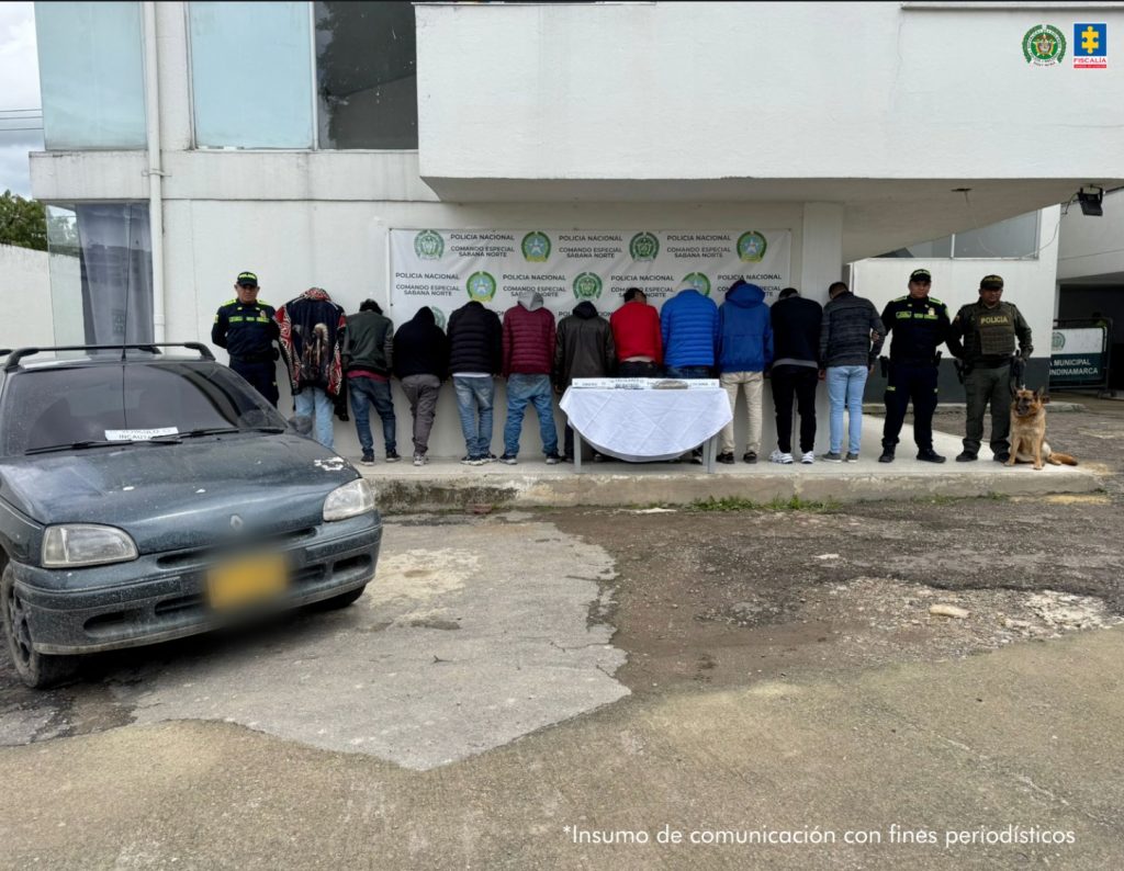 en la fotografia se pueden ver a los 11 detenidos de espaldas, en medio de 3 policias y un perro antinarcoticos, en frente de ellos hay un mesa con el material y estupefacientes incautados, tambien a un costado se puede ver un carro negro y de fondo hay un baking de la policia