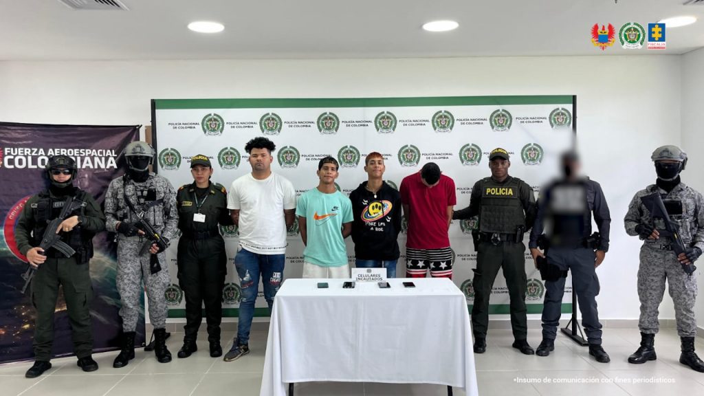 Los capturados aparecen custodiados por servidores de la Fiscalía y uniformados de la Policía y FAC, en la mesa algunos equipos de comunicación y de fondo, un pendón institucional.