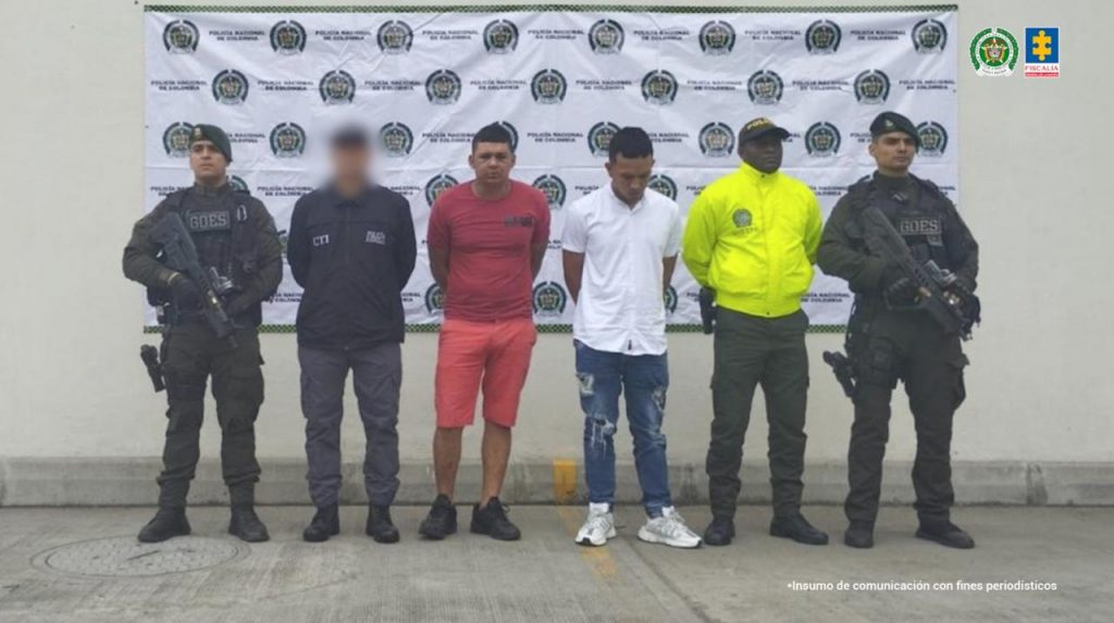 La imagen muestra a seis hombres alineados frente a una pared blanca con un cartel de fondo que tiene múltiples logos de la Policía Nacional de Colombia. A la izquierda y a la derecha de la imagen hay dos uniformados del GOES (Grupo de Operaciones Especiales), vestidos con trajes oscuros y armados con fusiles. En el centro de la imagen se encuentran dos hombres con ropa de civil, esposados y con la cabeza inclinada hacia abajo; uno lleva camiseta roja y pantalones cortos del mismo color, el otro viste camisa blanca y jeans rotos. A su lado están dos funcionarios más: uno con uniforme oscuro del CTI (Cuerpo Técnico de Investigación) con el rostro intencionalmente difuminado, y otro con chaqueta amarilla fluorescente de la Policía. En la parte superior derecha del cartel se observan los logos de la Policía Nacional y de la Fiscalía General de la Nación. La foto fue tomada con fines periodísticos y de comunicación institucional.