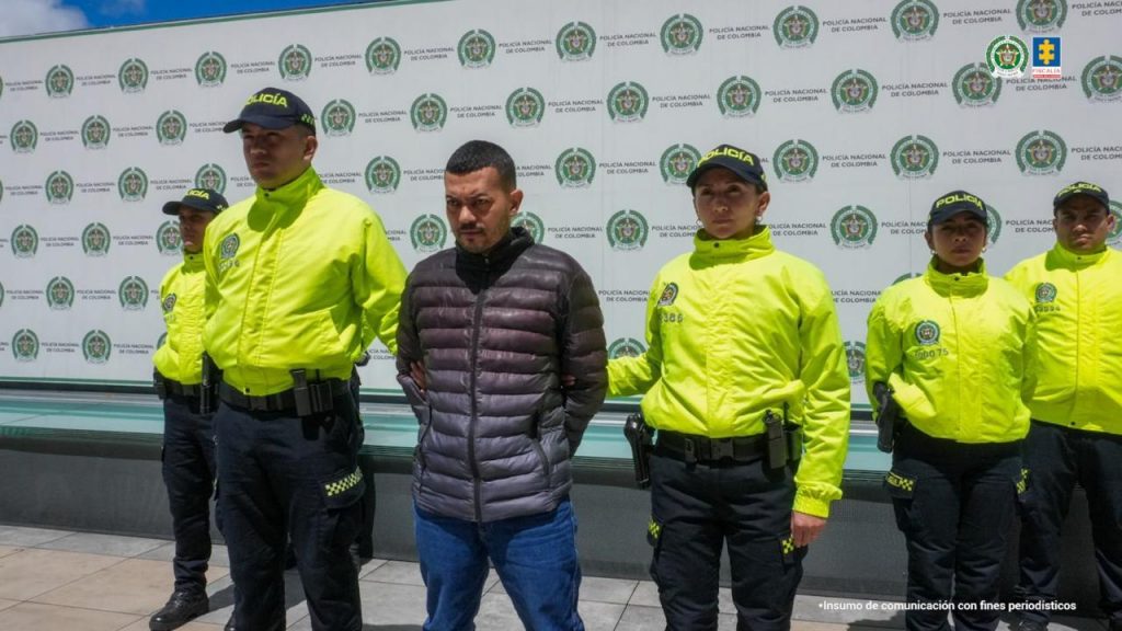 La imagen muestra a un hombre esposado en el centro, custodiado por varios policías uniformados. El hombre, de contextura media, lleva una chaqueta acolchada de color oscuro y jeans azules. Tiene el rostro serio y mira hacia el frente. Está custodiado a cada lado por policías vestidos con chaquetas amarillas fluorescentes y gorras negras con la palabra “POLICÍA” en letras mayúsculas. Al fondo, hay una pared cubierta con un pendón institucional repetido con el escudo de la Policía Nacional de Colombia. En la parte superior derecha de la imagen se observan los logos de la Policía Nacional y la Fiscalía General de la Nación.