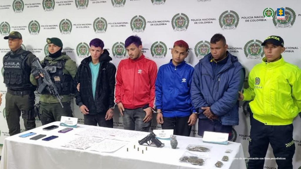 En la imagen aparecen cuatro procesados, en medio de una pareja de servidores de la Policía Nacional, más un uniformado al costado izquierdo. También, se observan los elementos incautados en diligencias de registro y allanamiento.