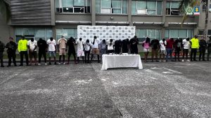 La imagen muestra una fila de personas detenidas, alineadas una al lado de la otra con la cabeza inclinada hacia abajo, aparentemente esposadas. Están frente a una mesa blanca cubierta con varios objetos (posiblemente evidencia), y un fondo institucional con logotipos repetidos, que parecen pertenecer a una entidad gubernamental o policial de Colombia. A los extremos de la fila hay varios agentes uniformados, algunos con chalecos reflectantes y otros con uniforme militar o policial. El lugar parece ser el exterior de una instalación oficial, posiblemente una estación de policía o edificio gubernamental. También se pueden ver los logotipos de la Fiscalía General de la Nación de Colombia y la Policía Nacional en la parte superior derecha de la imagen.  Preguntar a ChatGPT
