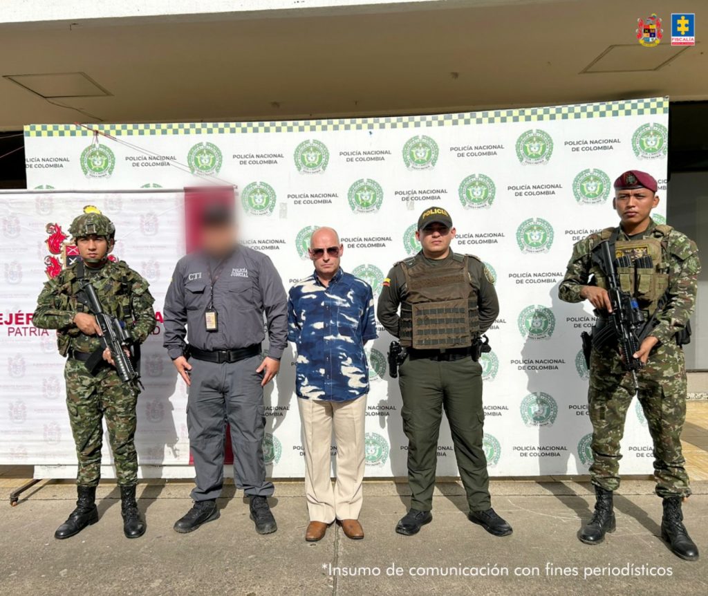 En la foto hay un militar en uniforme de camuflado con arma larga. Un funcionario del CTI (Cuerpo Técnico de Investigación) con el rostro intencionalmente difuminado, vestido con uniforme gris oscuro. Un hombre de civil (camisa tipo militar azul con blanco y pantalón claro), quien parece estar bajo custodia. Un uniformado de la Policía Nacional, con chaleco antibalas. Otro militar en uniforme de camuflado con boina burdeos, también con arma larga. Al fondo hay dos banners institucionales: uno con los logotipos de la Policía Nacional y otro del Ejército Nacional. En la esquina superior derecha aparecen los escudos de la Fiscalía General de la Nación y del Gobierno Nacional. En la parte inferior derecha se lee la advertencia: "Insumo de comunicación con fines periodísticos", lo que indica que la imagen fue difundida con fines informativos.