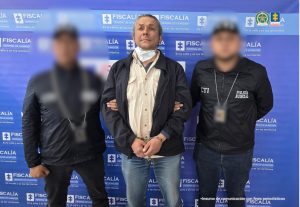 En la foto se observa a un hombre en el centro, esposado, escoltado por dos agentes de policía judicial (CTI) con los rostros difuminados. El detenido viste una camisa beige, chaqueta oscura y jeans, y lleva una mascarilla quirúrgica colgando del cuello. Los agentes a su lado visten ropa negra con las palabras "CTI" y "Policía Judicial" visibles en sus chaquetas.El fondo es un panel institucional azul con el logotipo de la Fiscalía General de la Nación de Colombia y su lema “En la calle y en los territorios”. También se aprecian los logotipos de otras entidades oficiales en la parte superior derecha.