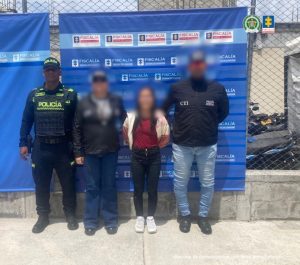 En la imagen se observa a cuatro personas de pie frente a un fondo institucional de la Fiscalía General de la Nación de Colombia, acompañado de logos oficiales. Dos de las personas en los extremos visten uniformes: a la izquierda, un policía con uniforme negro con detalles reflectivos verdes y el rótulo visible de "POLICÍA"; a la derecha, un agente del CTI (Cuerpo Técnico de Investigación) con chaqueta negra rotulada con "CTI" y "Policía Judicial".  En el centro están dos personas más, cuyas identidades han sido difuminadas (rostros borrosos), una de ellas aparentemente está siendo escoltada con las manos hacia atrás, lo que sugiere una posible captura o presentación ante medios judiciales. El fondo y la puesta en escena indican que la imagen forma parte de una comunicación oficial o judicial.  En la esquina superior derecha también aparecen los logos de Policía y de la Fiscalía. En la parte inferior derecha se alcanza a leer: "Insumo de comunicación con fines periodísticos".