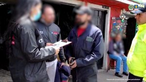 En la fotografía se observa un operativo de captura realizado por las autoridades colombianas. Cuatro personas están en primer plano: dos agentes del Cuerpo Técnico de Investigación (CTI) de la Fiscalía General de la Nación —una mujer y un hombre—, un agente de la Policía Nacional, y un hombre vestido con overol oscuro que parece estar siendo notificado o detenido.La funcionaria del CTI sostiene una carpeta con documentos, mientras lee o informa al capturado sobre el procedimiento. Todos los rostros están difuminados para proteger la identidad de los involucrados. Al fondo, se distingue la fachada de una vivienda con puerta metálica negra y paredes rojas, en lo que parece ser un barrio urbano. También se observa a otra persona sentada, desenfocada, al costado derecho. En la parte superior derecha aparecen los logotipos institucionales de la Fiscalía General de la Nación y la Policía Nacional de Colombia. En la esquina inferior derecha se lee: “Insumo de comunicación con fines periodísticos.”