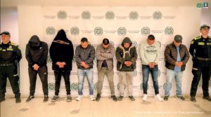 En la imagen se observa a ocho hombres en fila, seis de ellos en el centro con la cabeza inclinada hacia abajo y las manos juntas al frente, como si estuvieran esposados. Dos de las personas llevan sudaderas con capucha cubriéndose parcialmente el rostro.A los extremos, hay dos uniformados de la Policía Nacional de Colombia, vestidos con uniformes negros y chalecos reflectivos.  De fondo, se ve un telón con logotipos repetidos de la Policía Nacional de Colombia y la Fiscalía General de la Nación. En la esquina inferior derecha hay un texto que dice: “Insumo de comunicación con fines periodísticos”.