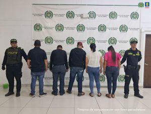 En la imagen se observa a seis personas de espaldas, alineadas en el centro, custodiadas por dos policías uniformados ubicados en los extremos izquierdo y derecho. Detrás de ellos hay un telón con el escudo y el nombre de la Policía Nacional de Colombia repetidos. En la parte superior derecha aparecen los logotipos de la Policía Nacional y de la Fiscalía General de la Nación. En la esquina inferior derecha se lee la frase “Insumo de comunicación con fines periodísticos”.
