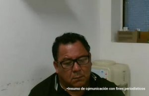 En la imagen se observa a un hombre de tez morena, con cabello corto y rizado de color oscuro, que lleva gafas y viste una camiseta tipo polo negra con ribetes blancos en el cuello. Está en un espacio interior de paredes blancas; detrás de él hay un dispensador de agua y, en una repisa o ventana, se ven algunas cajas y objetos pequeños. En la parte inferior de la imagen aparece el texto: "Insumo de comunicación con fines periodísticos".