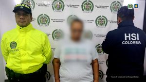 En la imagen se observa a tres personas frente a un fondo institucional de la Policía Nacional de Colombia.A la izquierda hay un hombre uniformado con chaqueta amarilla fluorescente y gorra que dice "POLICÍA".  En el centro hay una persona vestida con camiseta clara, cuya cara está desenfocada para proteger su identidad.  A la derecha hay otra persona de espaldas, con una chaqueta oscura que tiene las siglas "HSI COLOMBIA" en la parte trasera (lo que corresponde a Homeland Security Investigations de Estados Unidos).