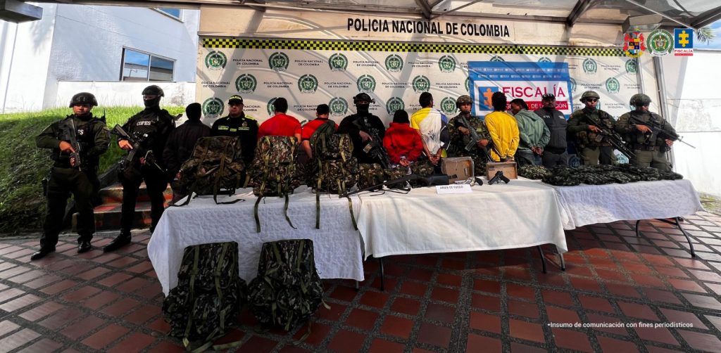 En la foto aparecen los detenidos de espalda acompañados de funcionarios de Cuerpo Técnico de Investigación (CTI), la Policía Nacional y el Ejército y al fondo pendones de las mismas instituciones.
