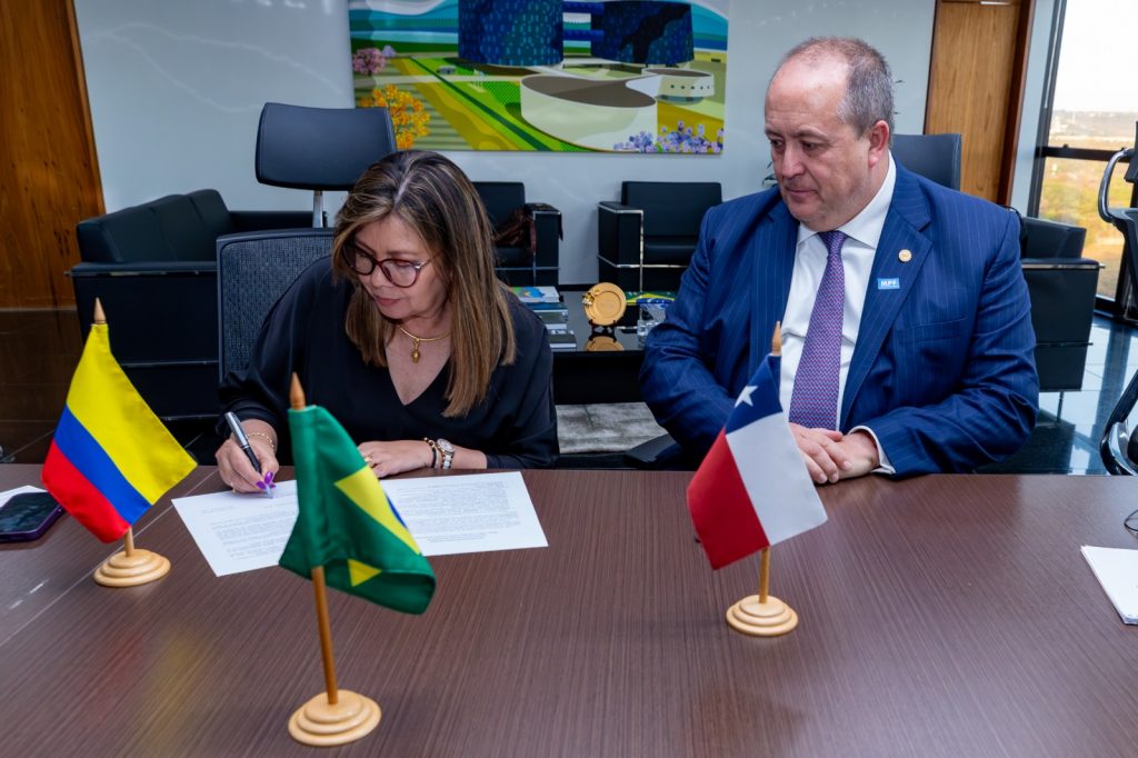 La fiscal general de la Nación, Luz Adriana Camargo Garzón, aparece sentada firmando un documento sobre una mesa. A su lado derecho está el fiscal nacional de Chile, Ángel Valencia Vásquez, quien observa el proceso. En la mesa se ven las banderas de Colombia, Brasil y Chile en pequeños soportes de madera. Al fondo, hay muebles de oficina y un cuadro colorido en la pared.