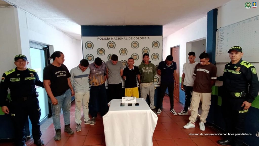 En la imagen se observan los nueve judicializados de frente y en medio de integrantes de la Policía Nacional. También, se visualizan los celulares incautados durante las diligencias de allanamiento.