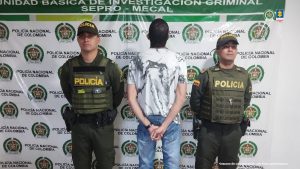 La imagen muestra a tres hombres frente a un fondo institucional de la Policía Nacional de Colombia.En el centro se encuentra un hombre de espaldas, con las manos juntas detrás, vistiendo una camiseta blanca con un diseño en tonos oscuros y un pantalón de mezclilla azul. A su lado izquierdo y derecho hay dos policías uniformados con chalecos antibalas que llevan la palabra "POLICÍA" en letras amarillas. Ambos están de frente a la cámara, en posición seria. El fondo tiene logotipos repetidos de la Policía Nacional de Colombia y en la parte superior se lee "UNIDAD BÁSICA DE INVESTIGACIÓN CRIMINAL SEPRO – MECAL".