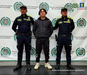 En la imagen se observan tres hombres de pie frente a un telón institucional de la Policía Nacional de Colombia.En el centro está un hombre vestido de negro, con chaqueta, pantalón deportivo y tenis claros. Tiene el cabello rizado y luce con las manos hacia los lados. A cada lado de él hay dos uniformados de la Policía Nacional, vestidos con uniforme oscuro con detalles en verde fosforescente. Ambos lo sujetan ligeramente por los brazos. En la parte superior del fondo aparecen los logos de la Policía Nacional de Colombia y, en una esquina, el de la Fiscalía General de la Nación. En la esquina inferior derecha se lee la frase: “Insumo de comunicación con fines periodísticos”.