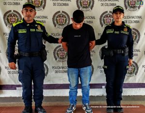 En la fotografía aparecen tres personas de pie frente a un telón institucional de la Policía Nacional de Colombia.En el centro está un hombre vestido con camiseta negra y pantalón de mezclilla azul, con la cabeza inclinada hacia abajo y las manos hacia atrás, en señal de estar bajo custodia. A su lado derecho e izquierdo se encuentran dos uniformados de la Policía Nacional, una mujer y un hombre, quienes lo sujetan de los brazos. Ambos visten uniforme azul oscuro con detalles en verde fosforescente y portan equipo de dotación policial. En el fondo se repite el escudo de la República de Colombia junto al texto “Policía Nacional de Colombia”. En la esquina superior derecha aparece el logotipo de la Fiscalía General de la Nación. En la parte inferior derecha está la frase: “Insumo de comunicación con fines periodísticos”.