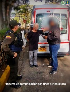 En la fotografía se observa a tres hombres en vía pública, frente a la parte trasera de un vehículo tipo furgón blanco con franjas rojas y placas amarillas.Uno de los hombres, de cabello canoso y ropa oscura, aparece con las manos hacia atrás mientras escucha a otro individuo con chaqueta negra y jeans azules que le muestra documentos. A la izquierda, un uniformado de la Policía Nacional, con chaleco y distintivos de la institución, observa la situación. En la parte superior derecha de la imagen aparecen los logotipos de la Policía Nacional y de la Fiscalía General de la Nación. Abajo, se lee la leyenda: “Insumo de comunicación con fines periodísticos”.