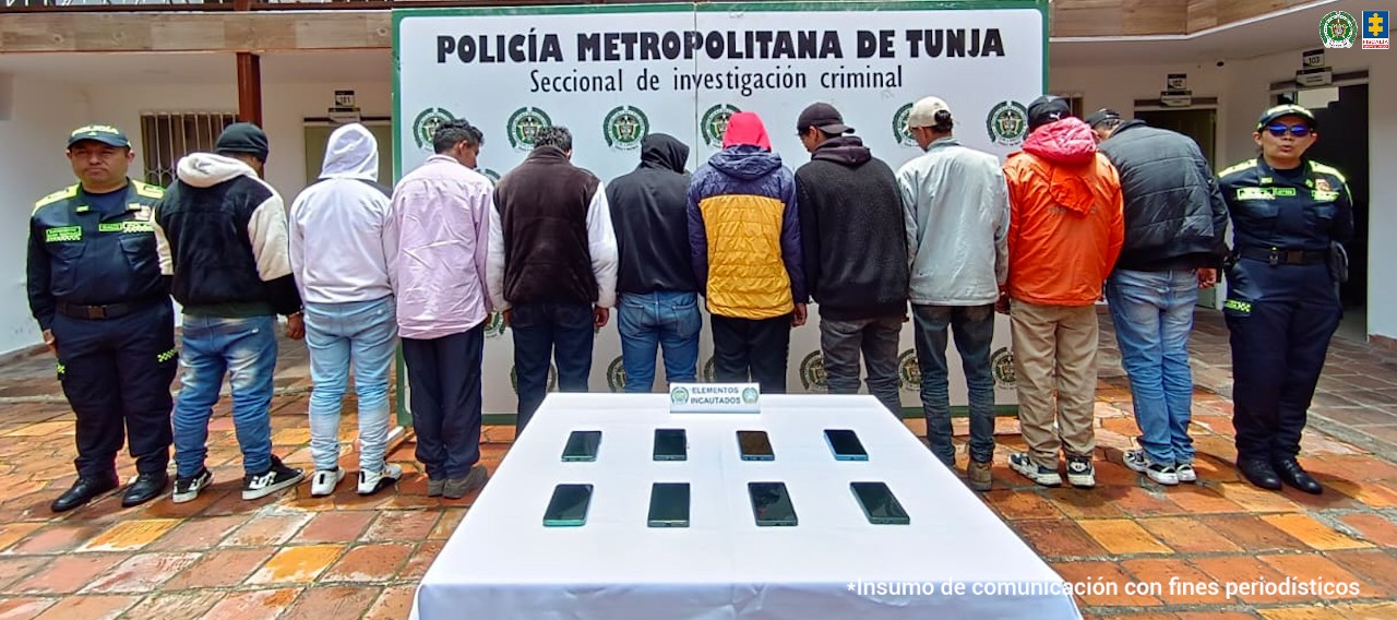 La imagen muestra a diez hombres de espaldas, alineados frente a un cartel que dice “Policía Metropolitana de Tunja – Seccional de investigación criminal”. A los lados de la fila hay dos uniformados de la Policía. En primer plano, sobre una mesa cubierta con un mantel blanco, se observan ocho teléfonos celulares incautados.