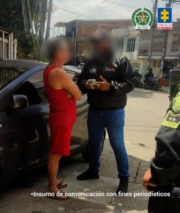 En la imagen se observa a dos hombres de pie en la vía pública, junto a un vehículo gris. Uno de ellos viste camiseta y pantaloneta rojas con sandalias, mientras que el otro lleva chaqueta negra con las siglas CTI y un pantalón de mezclilla azul, además sostiene documentos en sus manos.Al fondo se distinguen edificaciones de un barrio urbano y varias motocicletas estacionadas. En la parte superior derecha aparecen los logotipos de la Policía Nacional y de la Fiscalía General de la Nación. En la parte inferior de la imagen se lee la leyenda: “Insumo de comunicación con fines periodísticos”.