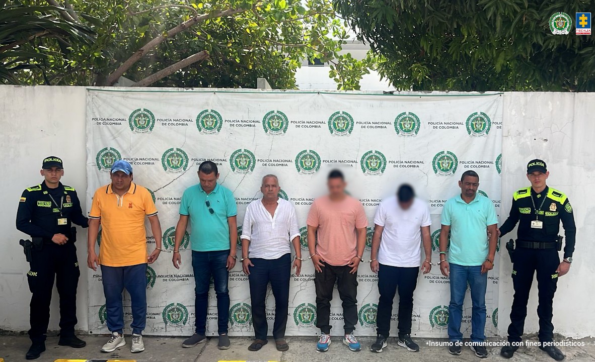 En la fotografía aparecen cinco hombres esposados y custodiados por dos policías uniformados. Los capturados están de pie frente a una pared blanca con un telón de la Policía Nacional de Colombia. La imagen fue tomada en un espacio exterior con árboles en la parte superior.