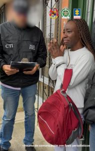 Una mujer con trenzas largas y camiseta clara aparece con gesto de angustia y las manos juntas frente a un funcionario del CTI de la Fiscalía, quien sostiene una carpeta y viste chaqueta negra con la palabra “Policía Judicial”. La mujer carga una mochila roja y se encuentra en la entrada de una vivienda, mientras otro agente la acompaña. En la parte superior se ven los logos institucionales de la Fiscalía y la Policía Nacional.