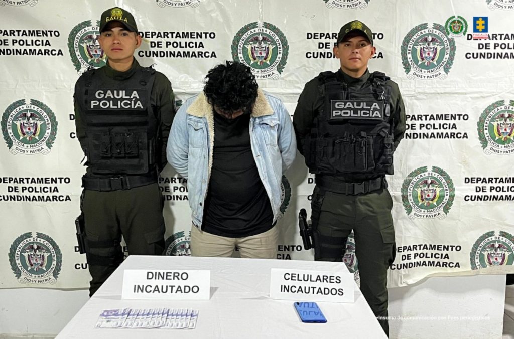 En la imagen se observa el procesado de frente y en medio de integrantes del Gaula de la Policía Nacional. También, se visualizan el celular y el dinero en efectivo incautado durante el procedimiento de captura. 