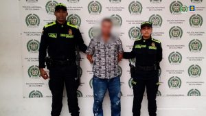 En la imagen se observa a dos uniformados de la Policía Nacional de Colombia, un hombre y una mujer, quienes escoltan a una persona en el centro de la fotografía. El individuo está vestido con camisa de manga corta estampada y pantalón de mezclilla azul, con las manos hacia atrás, en aparente condición de detenido.El fondo es un pendón institucional con los logos y emblemas de la Policía Nacional de Colombia; en la parte superior derecha también aparecen los logotipos de la Fiscalía General de la Nación y de la Policía Nacional.