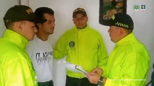 La imagen muestra a tres agentes de policía vestidos con chaquetas reflectivas de color amarillo neón y gorras negras con la palabra “Policía”. Están acompañando a un hombre vestido con camiseta blanca, quien parece estar bajo custodia. Uno de los policías sostiene unos documentos y parece estar leyéndolos o explicándolos al detenido.En la parte superior derecha de la imagen se observan dos logotipos institucionales: el de la Policía Nacional de Colombia y el de la Fiscalía General de la Nación. Abajo, en letras pequeñas, aparece un texto que dice: “Insumo de comunicación con fines periodísticos”.