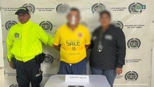 En la foto aparecen tres personas de pie frente a un fondo institucional con los logos de la Policía Nacional de Colombia y la Fiscalía General de la Nación.En el centro está un hombre con camiseta amarilla, que tiene el rostro difuminado, lo que indica que se trata de un capturado cuya identidad se protege.  A la izquierda se encuentra un policía uniformado con chaqueta amarilla de alta visibilidad y gorra negra con la palabra “POLICÍA”.  A la derecha hay otra persona con chaqueta oscura, también con el rostro difuminado, posiblemente un funcionario de apoyo en el procedimiento.  En primer plano, sobre una mesa, se observa un cartel que dice “EQUIPO MÓVIL” y un celular, aparentemente como evidencia incautada.