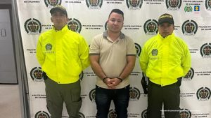 En la foto aparecen tres hombres.En el centro se ve a un hombre con una camiseta tipo polo clara, de manga corta, que tiene las manos juntas al frente y lleva esposas.  A cada lado de él hay dos policías vestidos con chaquetas amarillas de alta visibilidad y gorras negras con la palabra "POLICÍA".  El fondo es una lona blanca con el logotipo de la Policía Nacional de Colombia y también se observa el escudo de la Fiscalía General de la Nación.