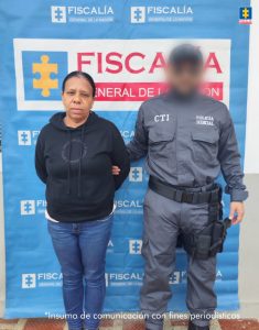 En la imagen se observa a una mujer escoltada por un funcionario del CTI de la Fiscalía General de la Nación. La mujer viste un buzo negro y un pantalón de mezclilla azul, mientras que el funcionario lleva uniforme gris con las insignias de “CTI” y “Policía Judicial”.Ambos posan frente a un pendón institucional de la Fiscalía General de la Nación, el cual muestra el logotipo de la entidad. En la parte inferior derecha de la imagen aparece una leyenda que indica: “Insumo de comunicación con fines periodísticos”.