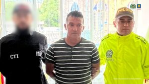 En la imagen se observa a tres hombres.El de en medio viste una camiseta a rayas horizontales en blanco y negro, con las manos hacia atrás, aparentemente esposado.  A su derecha (izquierda en la foto) está un hombre con uniforme negro que lleva las siglas CTI y "Policía Judicial"; su rostro está difuminado.  A su izquierda (derecha en la foto) hay un policía con uniforme fosforescente amarillo y gorra que dice POLICÍA.  En la parte superior derecha aparecen los logotipos de la Fiscalía General de la Nación de Colombia y de la Policía Nacional.