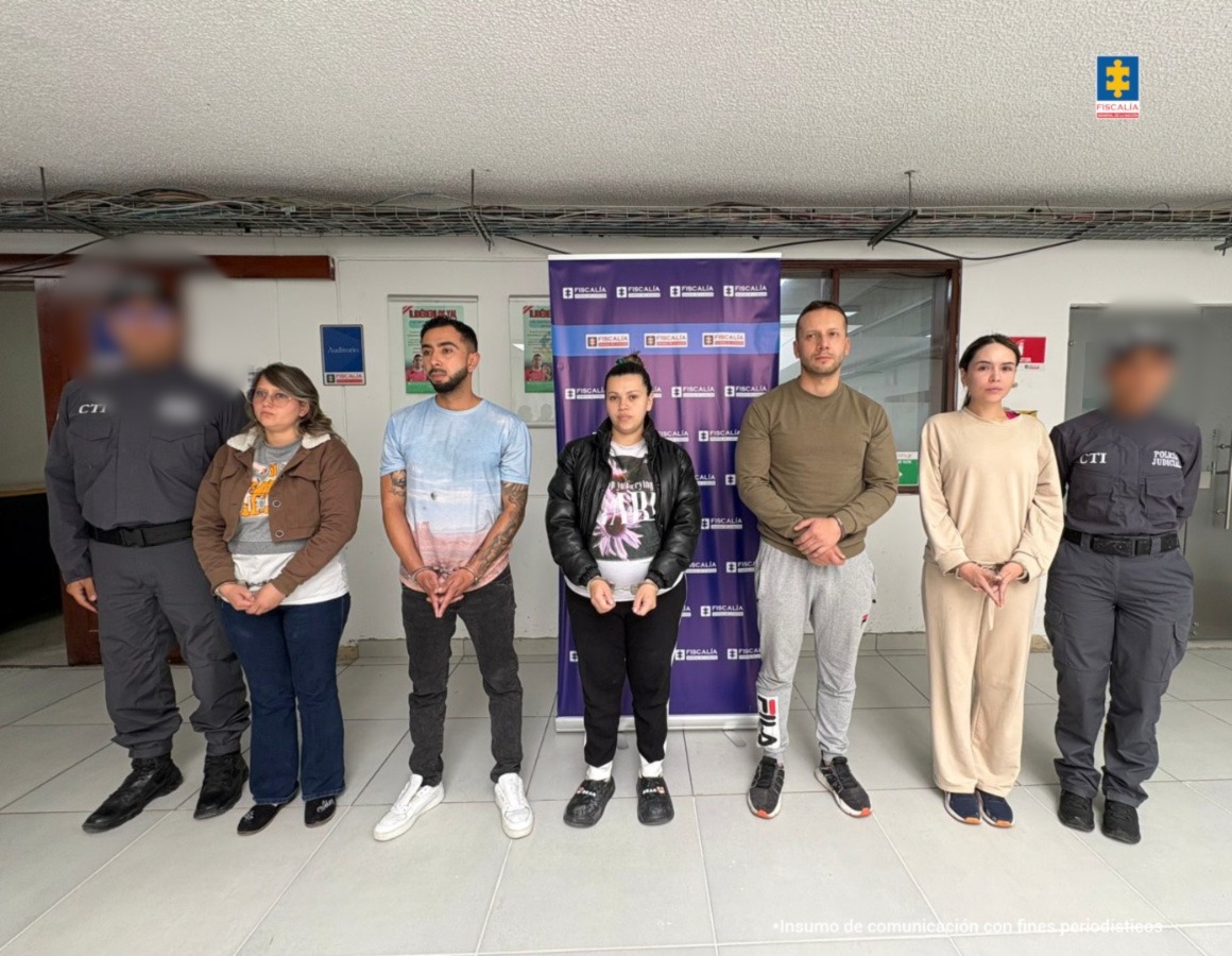 En la imagen se observan cinco personas capturadas, de pie, esposadas y custodiadas por dos agentes del CTI vestidos con uniformes oscuros. Están alineadas frente a un fondo institucional de la Fiscalía General de la Nación. Las personas visten ropa casual y deportiva.
