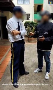 En la imagen se ve a dos hombres de pie conversando en lo que parece ser el patio o entrada de un conjunto residencial.Uno de ellos viste uniforme de vigilante, con pantalón azul oscuro con franjas amarillas, camisa blanca y gorra. El otro lleva chaqueta del CTI (Cuerpo Técnico de Investigación) con la inscripción “Policía Judicial” en la espalda y una gorra del mismo organismo. Sostiene un teléfono móvil en la mano. Ambos parecen estar en medio de una interacción formal o de verificación, probablemente relacionada con alguna diligencia. En la parte superior derecha se ve el logo de la Fiscalía General de la Nación de Colombia, y en la parte inferior hay un texto que dice: “Insumo de comunicación con fines periodísticos”.