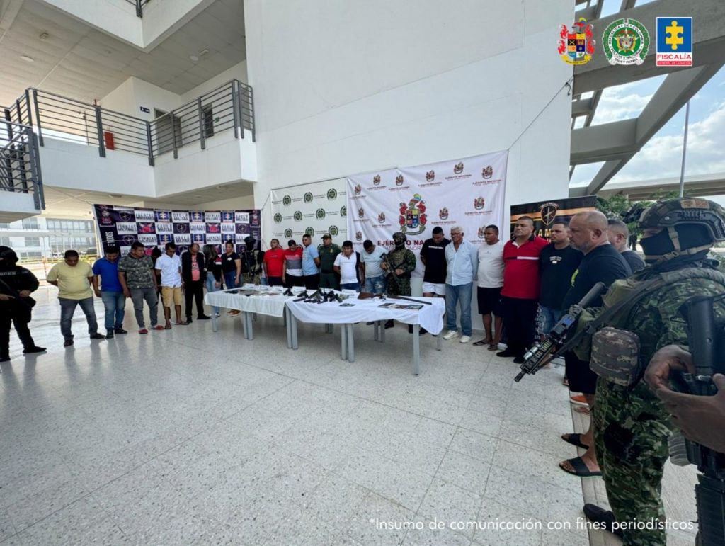 Fotografia en donde se pueden ver a los 19 capturados custodiados por uniformados del ejecitos nacional, detras de los capturados se pueden ver pendones de la policia y el ejercito, adelante de los capturados hay una mesa con elementos incautados