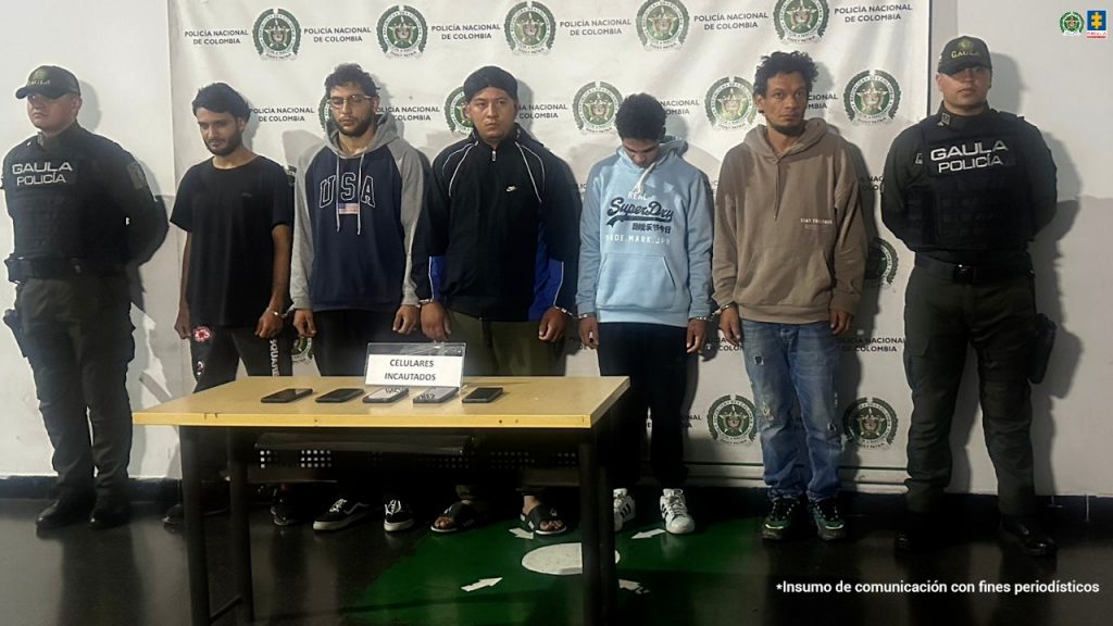 en la imagen se puede ver a 5 capturados en custiodio de dospolicias del Gaula, detras de ellos hay un pendon de la policia y en frente de estas personas hay una mesa con celulares incautados