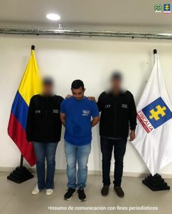 En la imagen se observa a tres hombres de pie, en un espacio interior. El hombre del centro viste una camiseta azul y jeans claros, y está acompañado a ambos lados por dos personas que llevan chaquetas negras con las siglas CTI y Policía Judicial. Detrás de ellos hay dos banderas: la de Colombia a la izquierda y la de la Fiscalía General de la Nación a la derecha. En la parte inferior de la imagen se lee la frase: “Insumo de comunicación con fines periodísticos”.