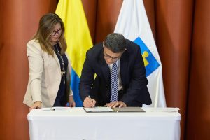 En la imagen se observa a dos personas en un entorno formal. Un hombre, vestido con traje oscuro y corbata, está firmando un documento sobre una mesa cubierta con un mantel blanco. A su lado, una mujer con gafas, chaqueta beige y blusa oscura lo acompaña mientras observa la firma.Detrás de ellos se aprecian dos banderas: una con los colores amarillo, azul y rojo (la bandera de Colombia) y otra con un símbolo institucional en blanco y azul. El ambiente sugiere un acto oficial o protocolario, posiblemente relacionado con la firma de un acuerdo o documento gubernamental.