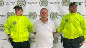 En la imagen se observa a tres hombres de pie frente a un fondo institucional con los logotipos de la Policía Nacional de Colombia.En el centro está un hombre de cabello canoso y bigote, vestido con una camisa tipo polo blanca, con las manos hacia atrás, aparentemente esposadas. A cada lado se encuentran dos uniformados de la Policía Nacional, ambos con chaquetas reflectivas de color amarillo neón, gorras negras con la palabra “POLICÍA”, y portando su dotación de servicio. En la parte superior derecha de la imagen aparecen los logotipos de la Policía Nacional y de la Fiscalía General de la Nación, y en la parte inferior se lee la frase:
“Insumo de comunicación con fines periodísticos”.