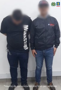 En la imagen se observan dos hombres de pie frente a una pared blanca, ambos con el rostro difuminado para proteger su identidad.La persona ubicada a la izquierda viste camiseta negra con la palabra “MONASTRY” en letras blancas verticales, pantalón oscuro y zapatos negros. A la derecha se encuentra otro hombre que lleva una chaqueta negra con logotipos institucionales, pantalón de mezclilla azul y zapatos marrones. En la parte superior derecha aparecen los logotipos de la Fiscalía General de la Nación y de la Policía Nacional de Colombia, y en la parte inferior de la imagen se lee la frase:
“Insumo de comunicación con fines periodísticos”.