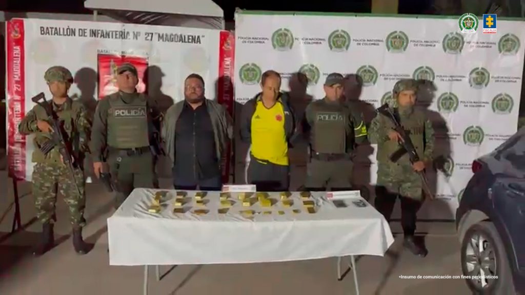 En la imagen se observa a dos hombres capturados de pie, escoltados por varios uniformados del Ejército y la Policía Nacional. Frente a ellos hay una mesa cubierta con un mantel blanco, sobre la cual reposan numerosos lingotes de oro organizados en filas. Al fondo se distinguen pendones institucionales del Batallón de Infantería N.º 27 Magdalena, la Policía Nacional y la Fiscalía General de la Nación.