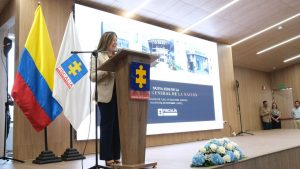 En la foro aparece la fiscal Luz Adriana Camargo vestida con chaqueta beige, pantalon oscuro. Adelante de ella está un atril con el logo de la Fiscalía. En la parte de atrás se ve una bandera blanca con el logo de la Fiscalía.